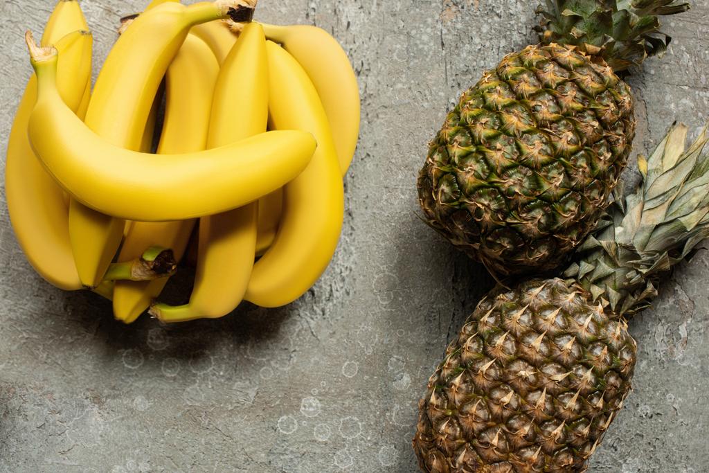 top view of colorful delicious bananas and pineapples on grey concrete surface - Photo, Image