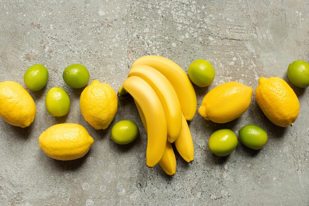 top view of colorful bananas, limes and lemons on grey concrete surface - Photo, Image