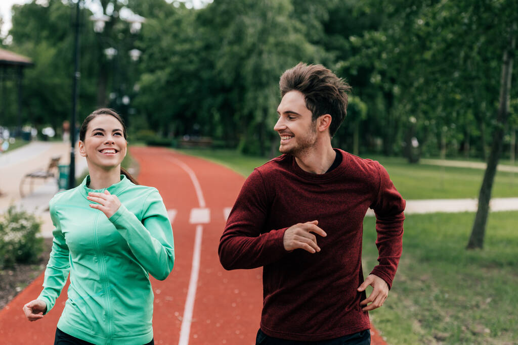 uśmiechnięty mężczyzna patrząc na pozytywną dziewczynę podczas joggingu w parku  - Zdjęcie, obraz