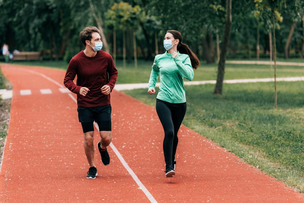 Бегуны в медицинских масках, смотрящие друг на друга во время тренировок на беговой дорожке в парке
  - Фото, изображение