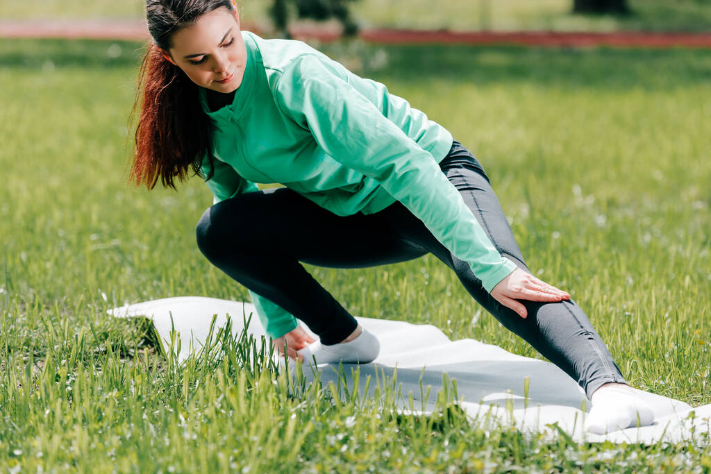 Selective focus of sportswoman warming up on fitness mat in park  - Photo, Image