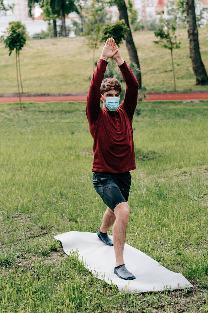 Sportman in medisch masker met opgeheven handen training op fitness mat in park  - Foto, afbeelding