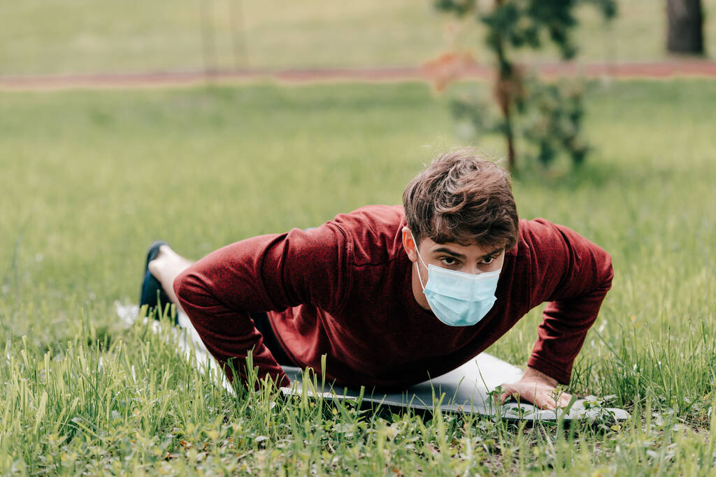 Selektiver Fokus des Sportlers mit medizinischer Maske beim Liegestützen auf der Fitnessmatte im Park  - Foto, Bild