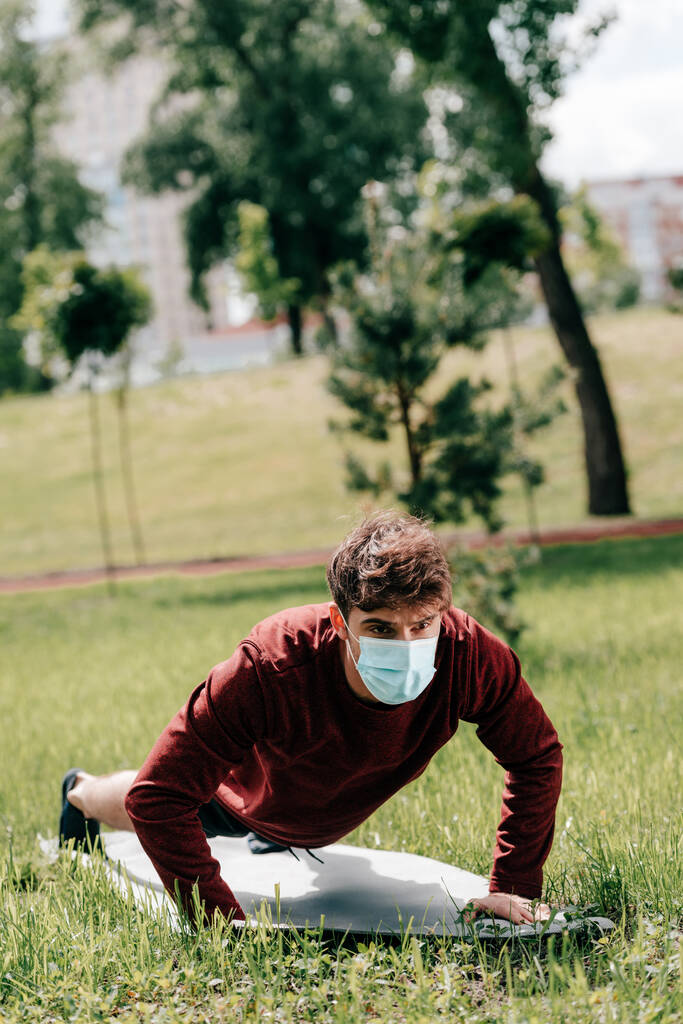 Enfoque selectivo del joven deportista en máscara médica haciendo flexiones en la estera de fitness en la hierba en el parque
  - Foto, Imagen