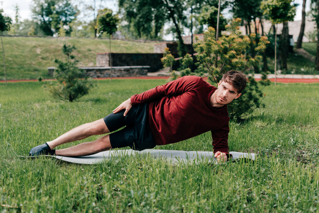 Enfoque selectivo de deportista guapo haciendo tablón lateral mientras entrena en la estera de fitness en el parque
  - Foto, Imagen