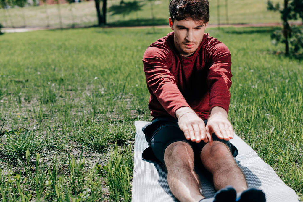 Selective focus of handsome man stretching on fitness mat on grass in park  - Photo, Image