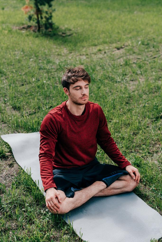 Hombre guapo con los ojos cerrados sentado en postura de yoga en la estera de fitness en el parque
  - Foto, imagen