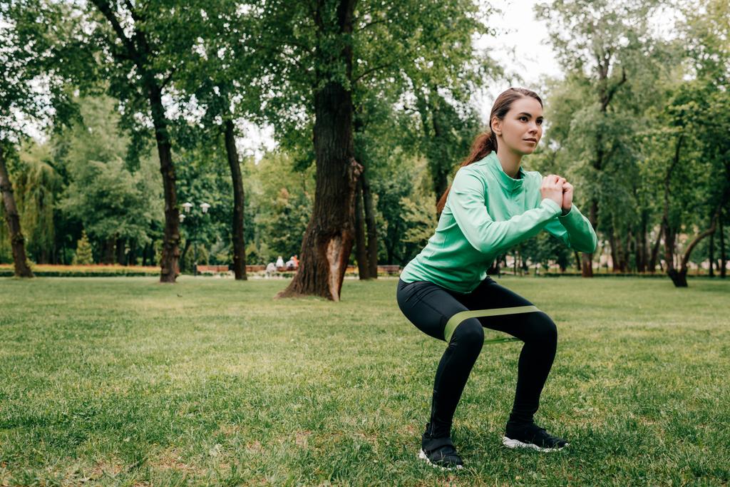 Aantrekkelijke sportvrouw doet squat met verzetsband in park  - Foto, afbeelding