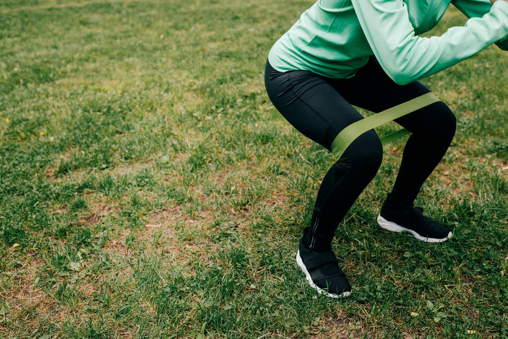 Cropped view of sportswoman doing squat with resistance band on grass in park  - Photo, Image