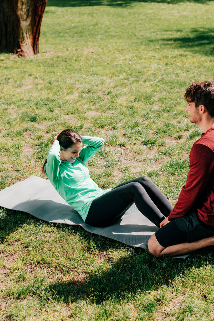 Smiling sportswoman doing abs near boyfriend on fitness mat in park  - Photo, Image