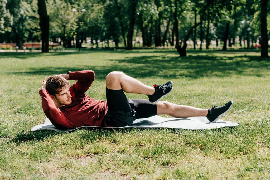 Vista lateral del deportista guapo haciendo abdominales en la alfombra de fitness en el parque
  - Foto, Imagen