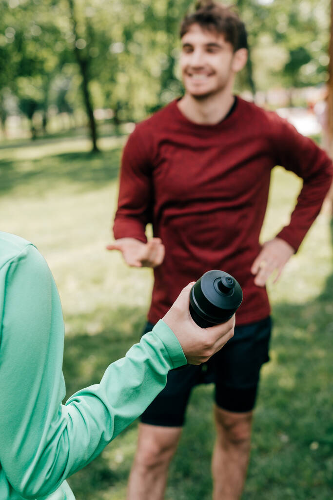 公園内の笑顔のスポーツマンの近くにスポーツボトルを保持するスポーツ女性の選択的な焦点  - 写真・画像