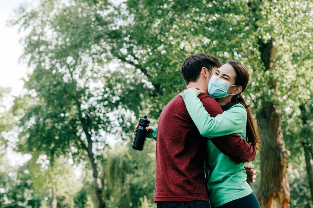 sportivo abbracciare fidanzata in maschera medica tenendo bottiglia sportiva nel parco
  - Foto, immagini