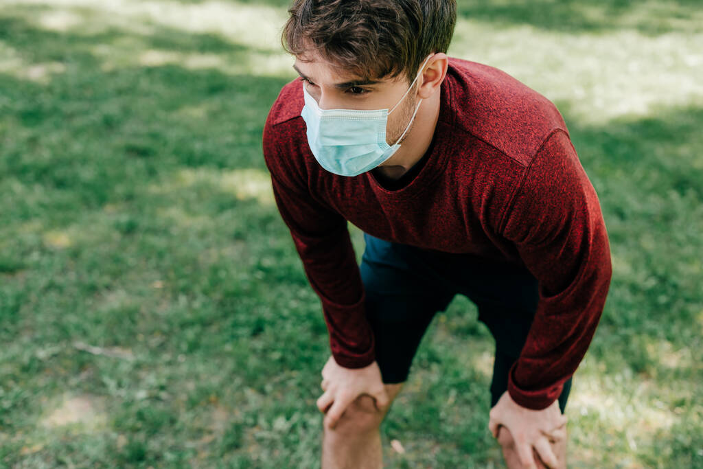 Νεαρός αθλητής με ιατρική μάσκα στέκεται στο πάρκο  - Φωτογραφία, εικόνα