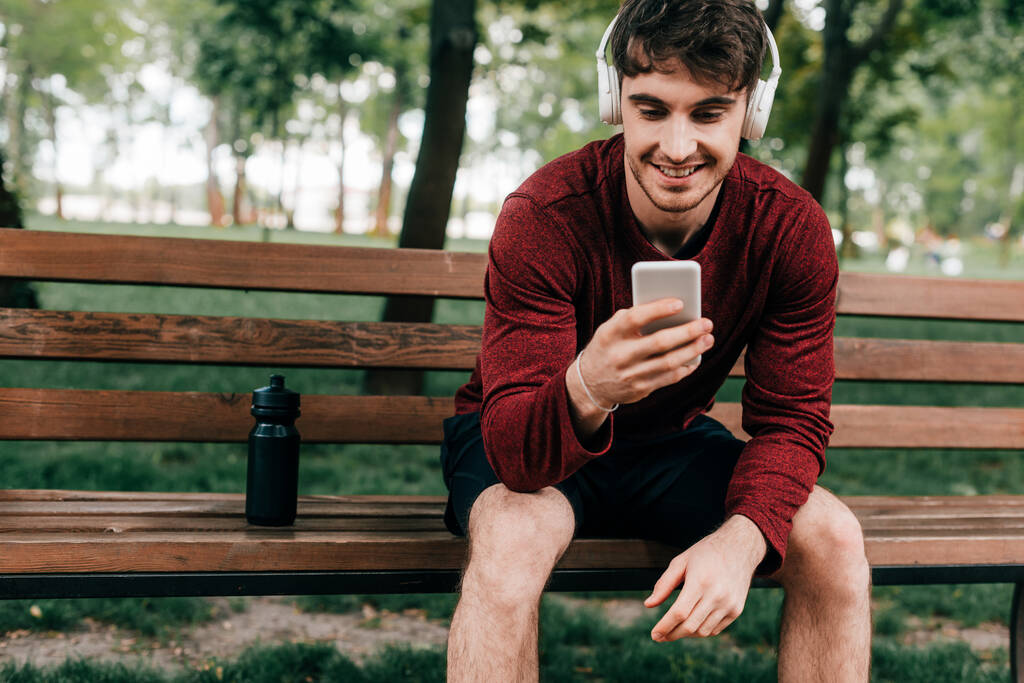 Uśmiechnięty sportowiec w słuchawkach za pomocą smartfona w pobliżu butelek sportowych na ławce w parku  - Zdjęcie, obraz