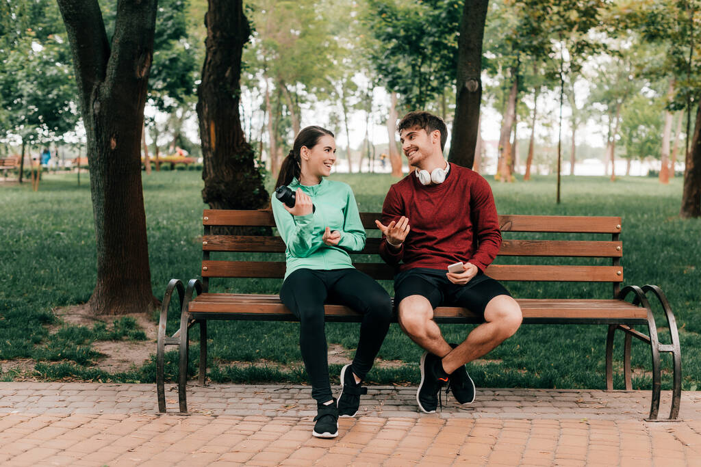 Sportif souriant dans les écouteurs regardant petite amie avec bouteille de sport sur le banc dans le parc  - Photo, image
