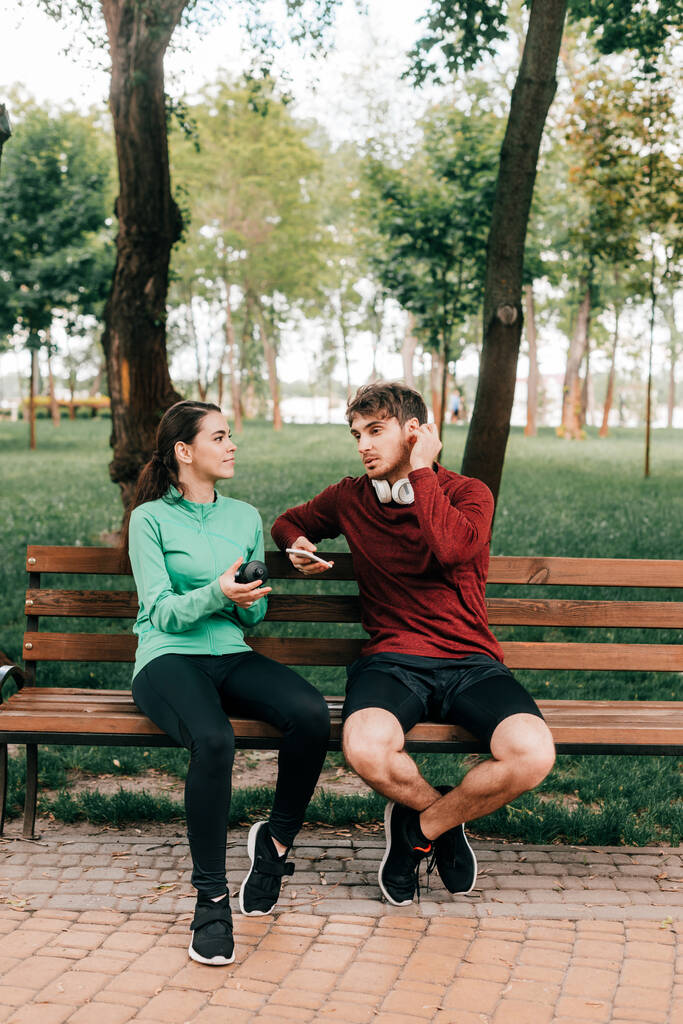 Улыбающаяся спортсменка держит спортивную бутылку возле бойфренда в наушниках со смартфоном на скамейке в парке
  - Фото, изображение