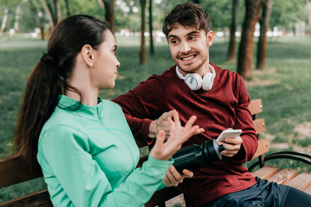 Selektivní zaměření usměvavý sportovec ve sluchátkách držící smartphone při rozhovoru s přítelkyní na lavičce v parku  - Fotografie, Obrázek