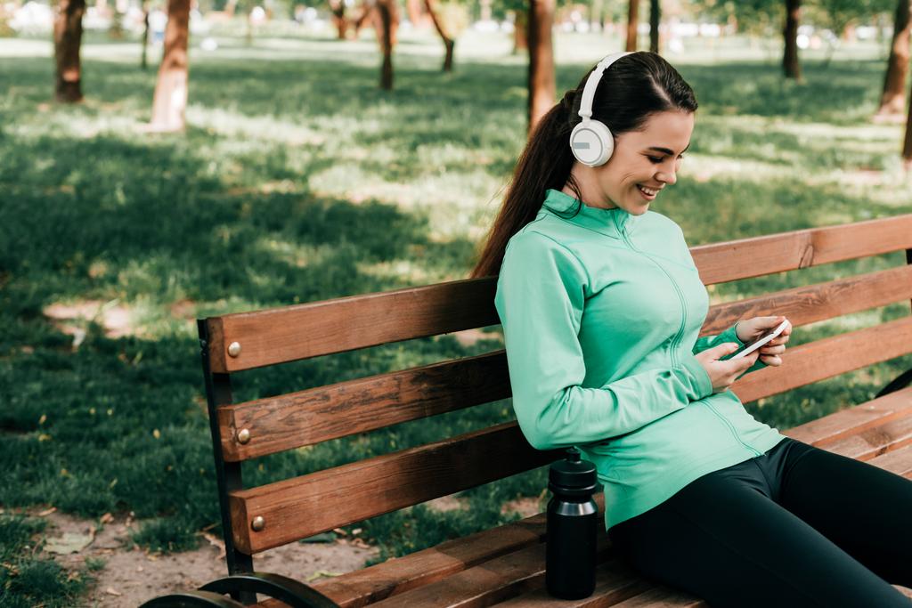 Sportiva sorridente in cuffia utilizzando smartphone vicino a bottiglia sportiva su panchina nel parco
  - Foto, immagini