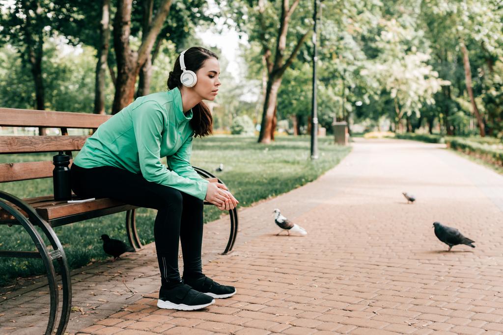 Spor kulaklıklı kadın spor şişesinin yanında oturuyor ve parkta bankta akıllı telefon var.  - Fotoğraf, Görsel