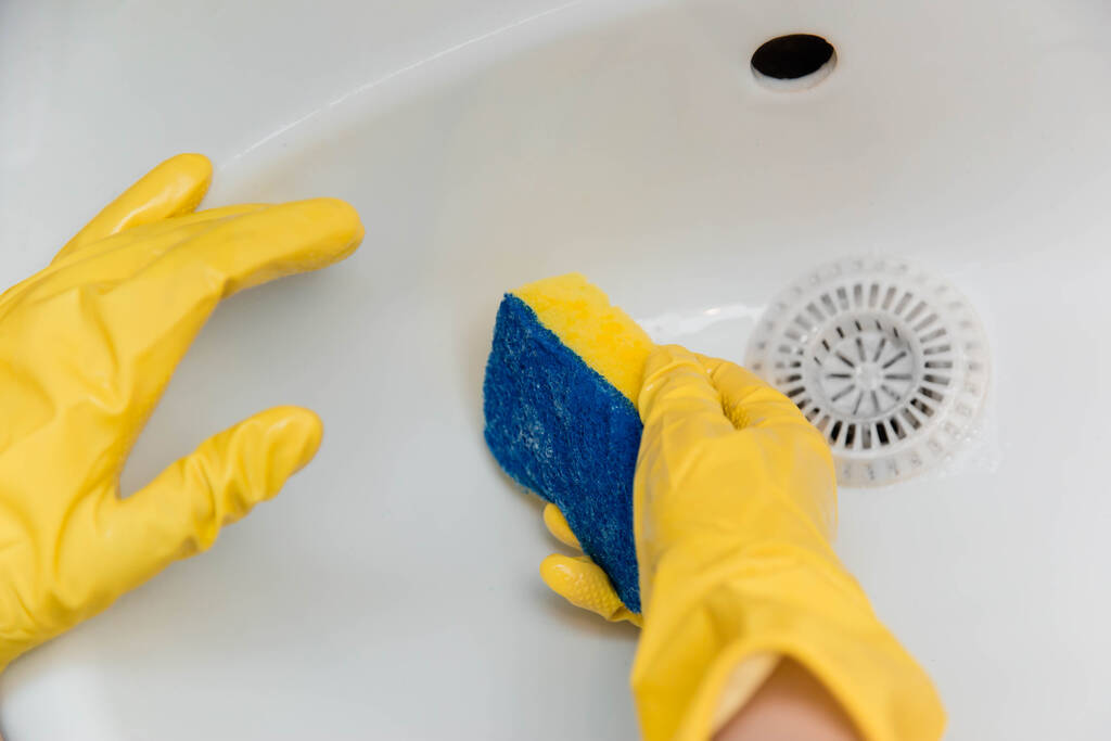 Close-up of a hand in a yellow rubber glove wipes a white sink with a sponge. - Photo, Image