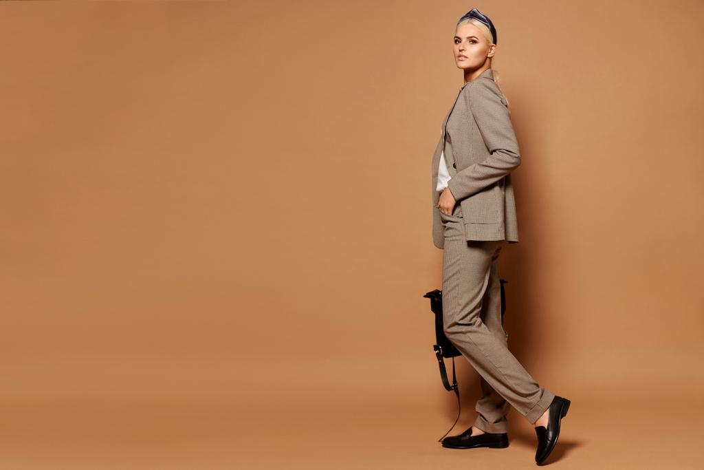 Vista lateral de la joven mujer de negocios hermosa en un pantalón y chaqueta aislada sobre un fondo beige. Retrato de cuerpo entero. Copiar espacio
 - Foto, imagen
