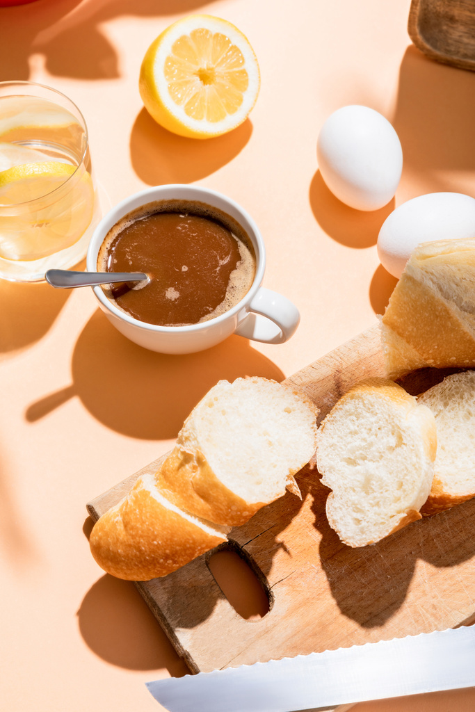 tazza di caffè con cucchiaino, uova, baguette e bicchiere d'acqua con limone per colazione sul tavolo beige - Foto, immagini