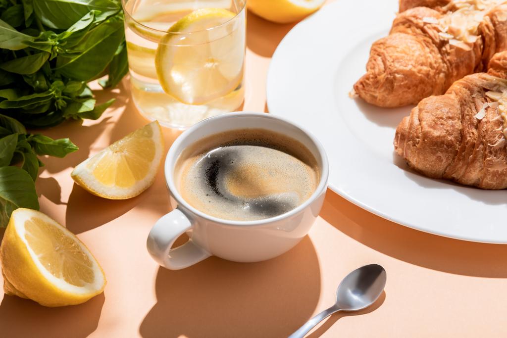 káva, croissanty a sklenice vody s citronem k snídani na béžovém stole - Fotografie, Obrázek