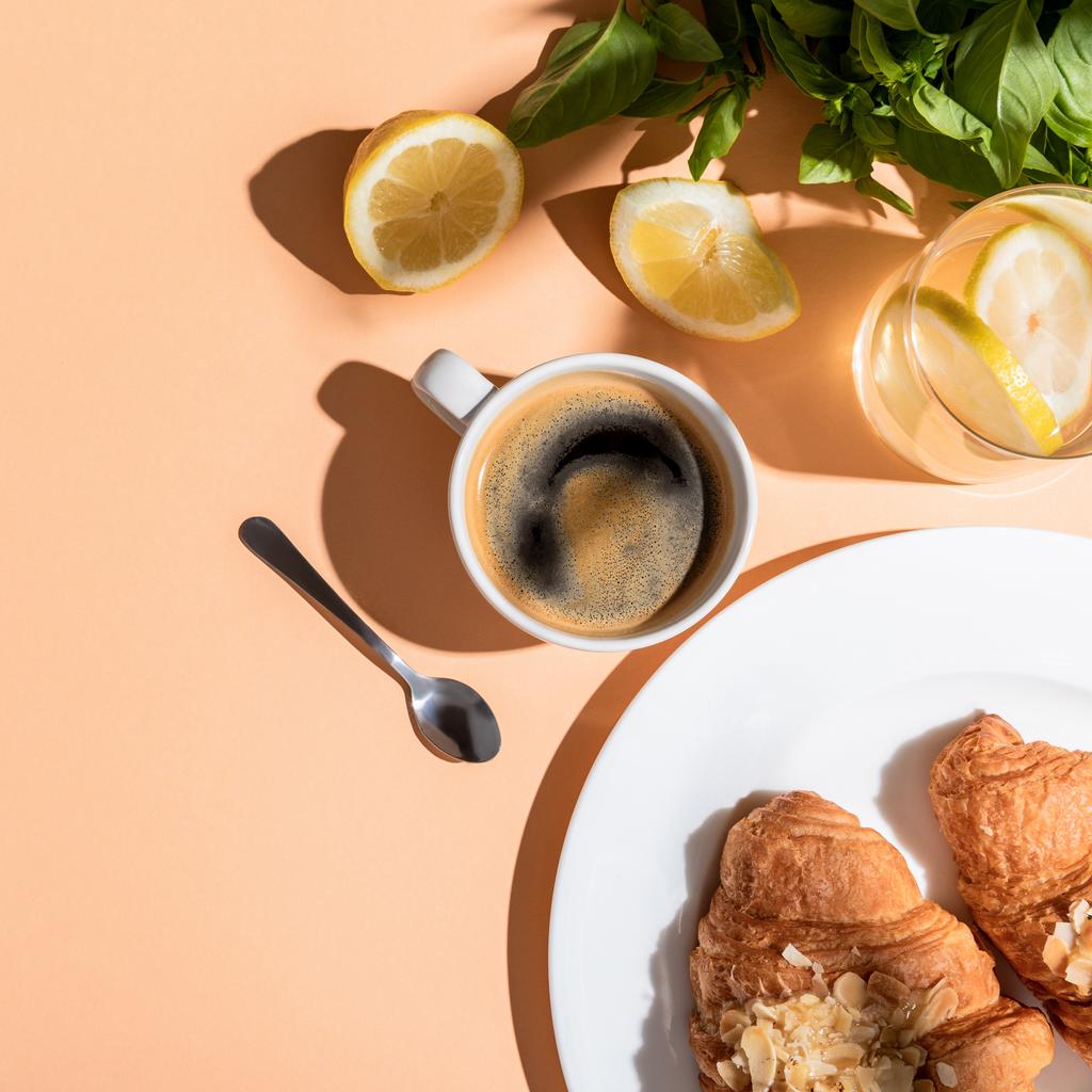 vista superior de la taza de café, vegetación, limones y croissants para el desayuno en la mesa beige
 - Foto, Imagen