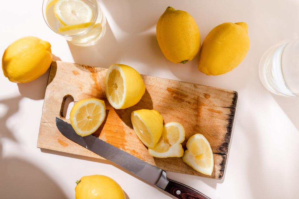 vrchní pohled na celé a broušené citrony na dřevěné desce s nožem a sklenicemi vody na šedém stole - Fotografie, Obrázek