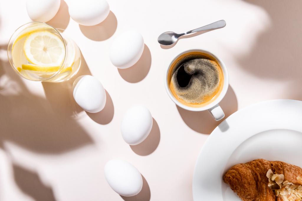 top view of chicken eggs, water with lemon, coffee cup and croissant for breakfast on grey table with shadows - Photo, Image