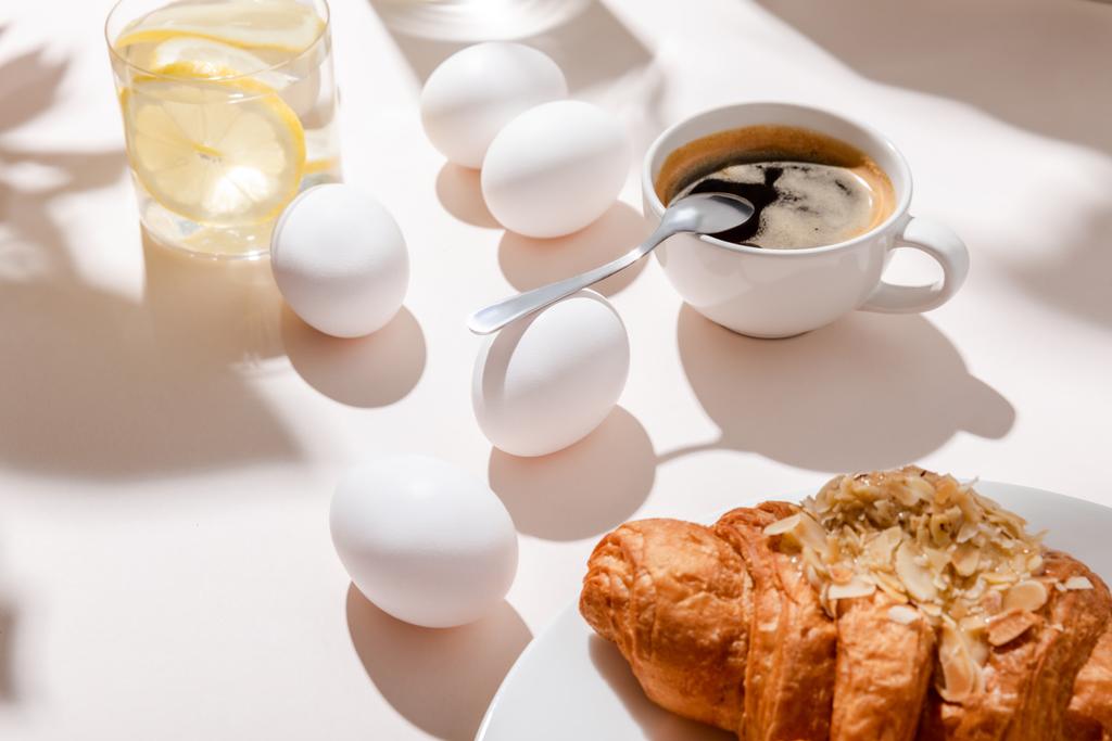 kuřecí vejce, croissant, šálek kávy a sklenice vody s citronem k snídani na šedém stole se stíny - Fotografie, Obrázek