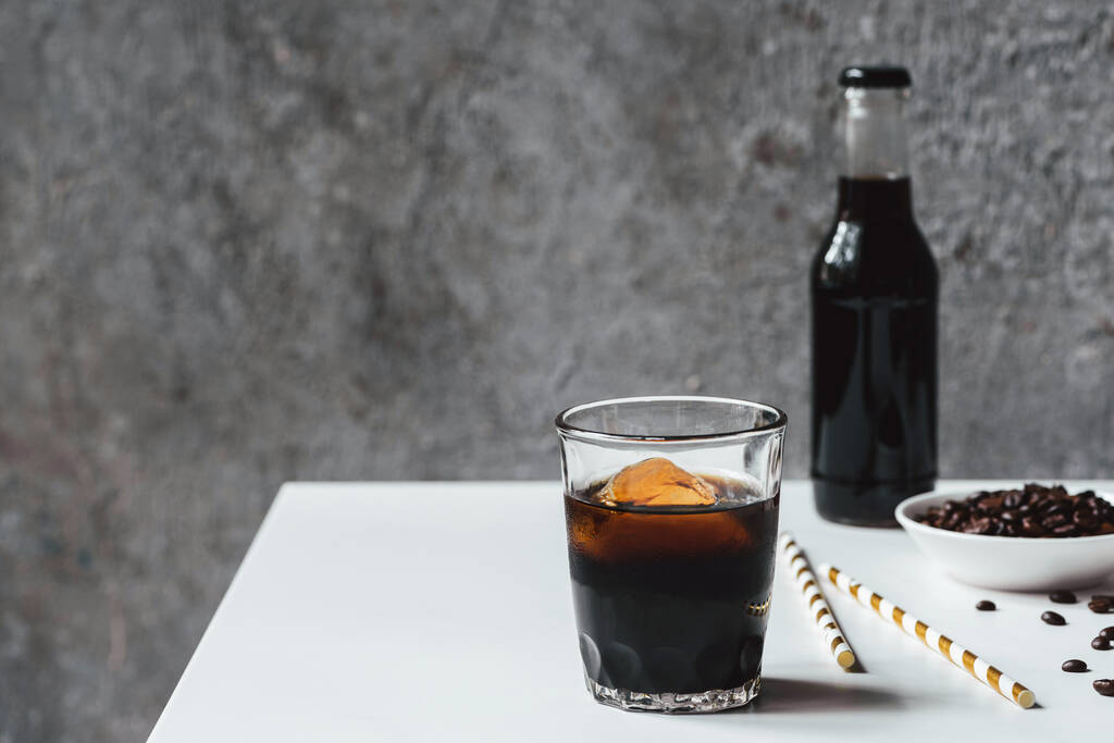 selective focus of cold brew coffee with ice in glass and bottle near drinking straws and coffee beans on white table - Valokuva, kuva