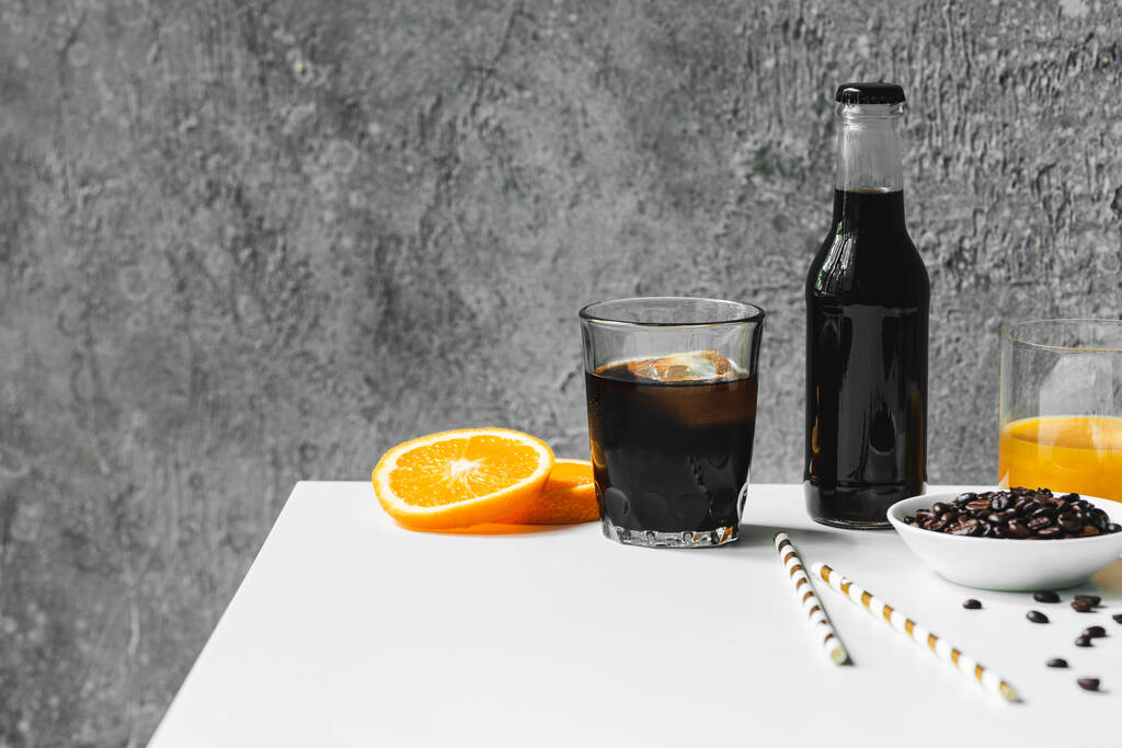 селективный фокус холодного кофе со льдом в стакане и бутылкой рядом с апельсиновым соком, питьевой соломы и кофейных зерен на белом столе
 - Фото, изображение