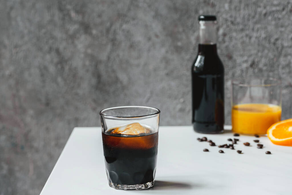foco seletivo de café frio brew com gelo em vidro e garrafa perto de suco de laranja e grãos de café na mesa branca
 - Foto, Imagem