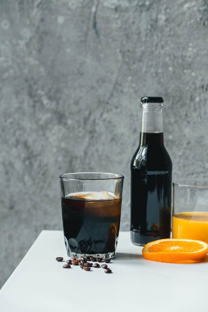 Холодный кофе со льдом в стакане и бутылка возле апельсинового сока и кофейных зерен на белом столе
 - Фото, изображение