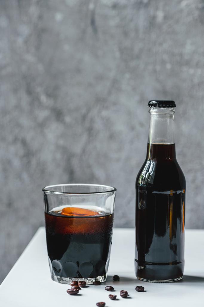 Холодный кофе со льдом в стекле и бутылка возле кофейных зерен на белом столе
 - Фото, изображение