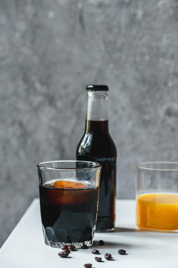messa a fuoco selettiva di caffè freddo birra con ghiaccio in vetro e bottiglia vicino succo d'arancia e chicchi di caffè sul tavolo bianco
 - Foto, immagini