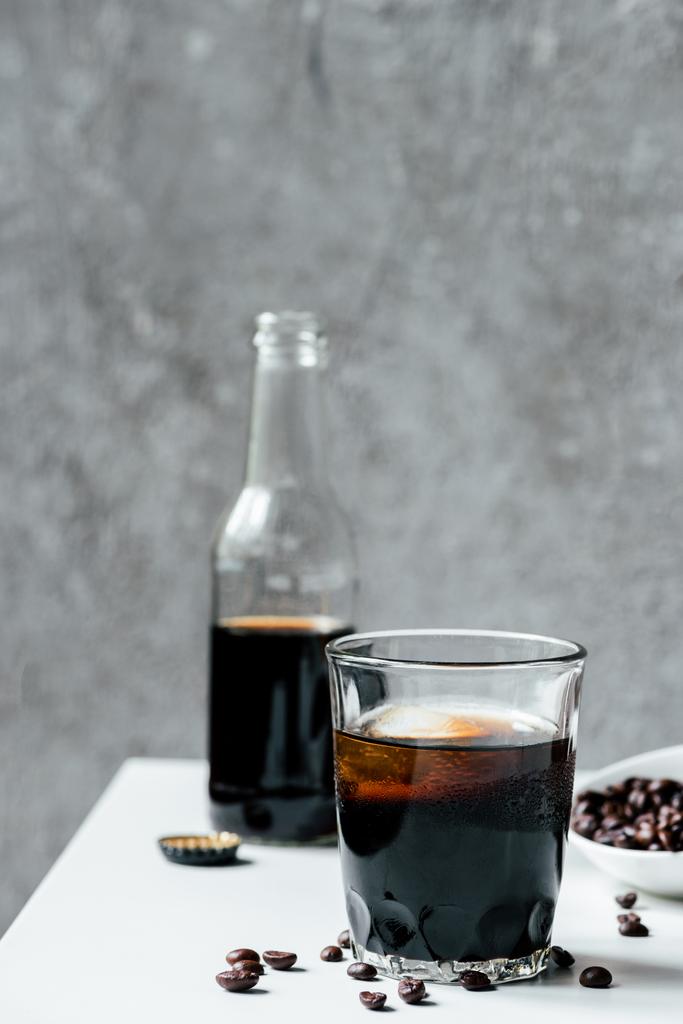 selektiver Fokus von kaltem Brühkaffee mit Eis im Glas in der Nähe der Flasche und Kaffeebohnen auf weißem Tisch - Foto, Bild