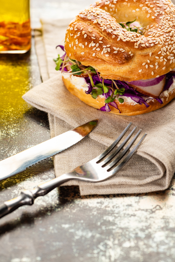fresh delicious bagel with meat, red onion, cream cheese and sprouts on napkin with cutlery on textured surface - Photo, Image