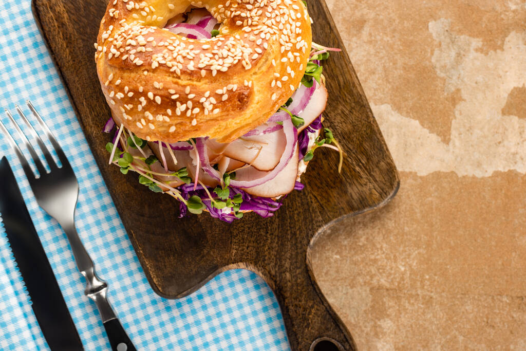 top view of fresh delicious bagel with meat, red onion and sprouts on wooden cutting board and plaid blue napkin with cutlery - Photo, Image