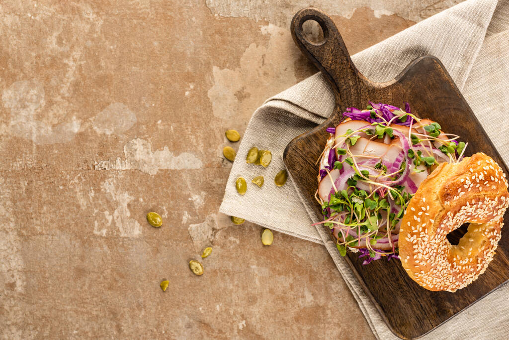 vista superior del delicioso bagel fresco con carne, cebolla roja y brotes en una tabla de cortar de madera en una servilleta con semillas de calabaza en una superficie beige envejecida
 - Foto, imagen