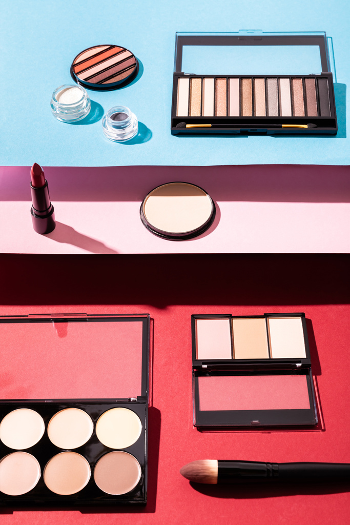 Rouge und Lidschattenpaletten in der Nähe von Kosmetikbürsten, Lippenstift und Gesichtspuder auf Karminrot, Rosa und Blau - Foto, Bild
