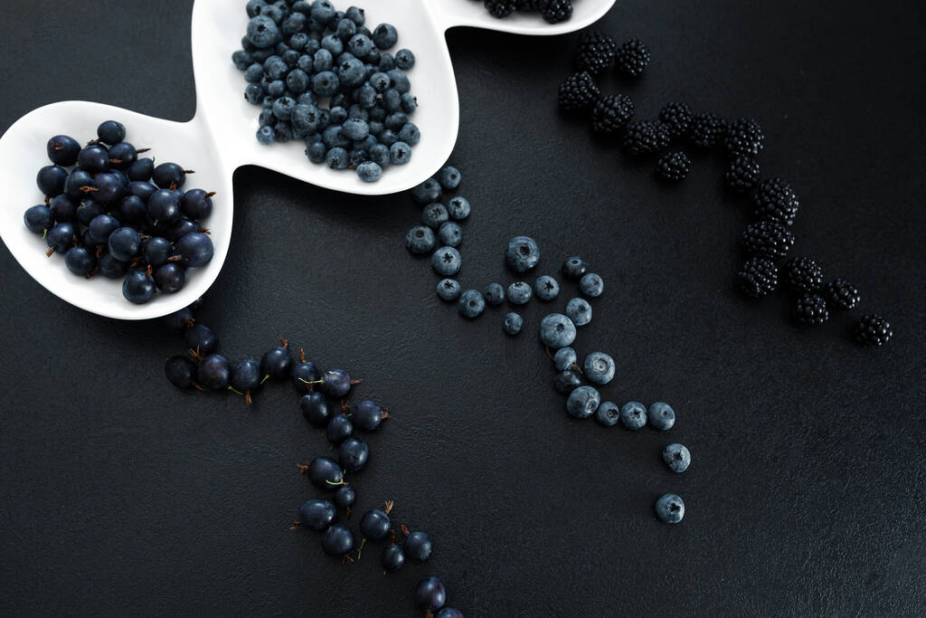 Set verschiedener schwarzer Beeren in einem weißen Teller auf einem schwarzen Tisch. Stilvolle saisonale Vitamine. - Foto, Bild
