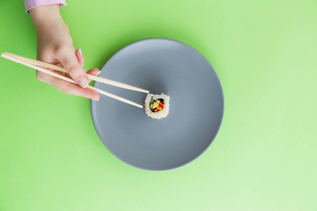 Płaskie ułożenie żeńskiej dłoni trzymającej pałeczki i jedna rolka sushi na płycie izolowanej na zielonym tle. - Zdjęcie, obraz