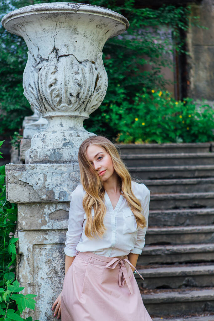 Portrait of a beautiful blond girl standing next to an old building with a decorative antique vase and stairs - Photo, Image