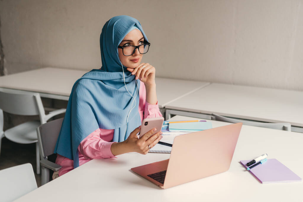 joven mujer musulmana bastante moderna en hijab trabajando en el ordenador portátil en la sala de oficina, educación en línea, freelancer trabajo remoto, usando el pensamiento smatphone
 - Foto, Imagen