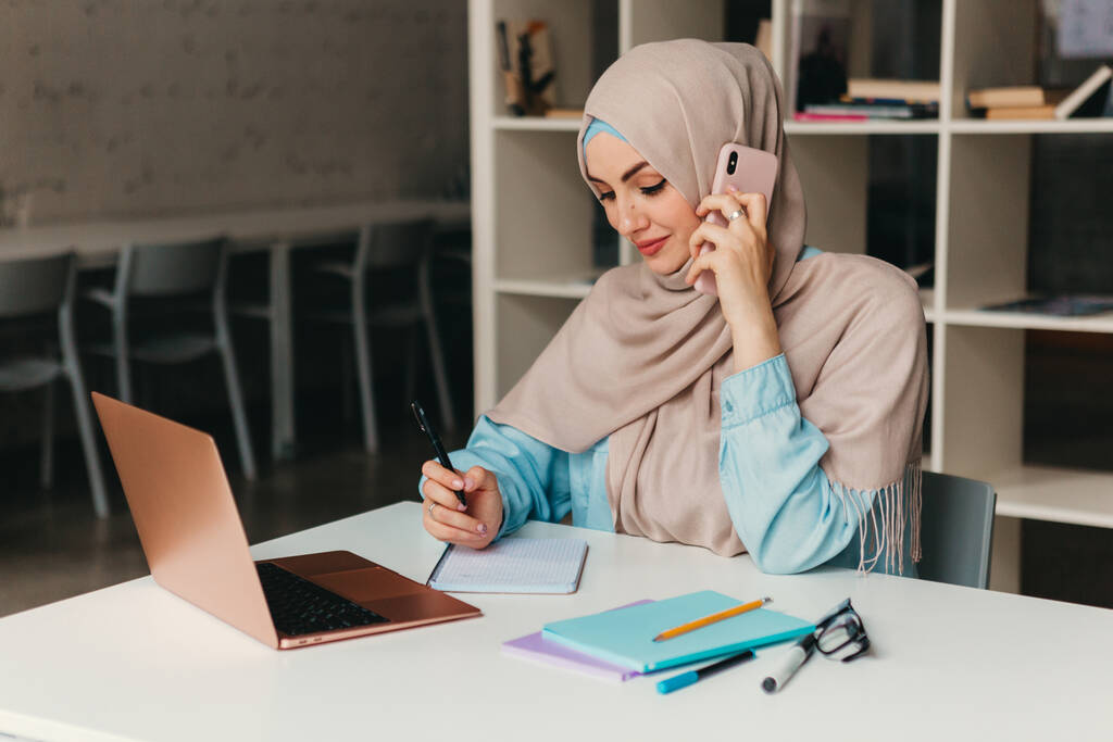fiatal meglehetősen modern muszlim nő hidzsáb dolgozik laptop irodai szobában, oktatás online, távoli munka szabadúszó, beszél okostelefon jegyzeteket - Fotó, kép