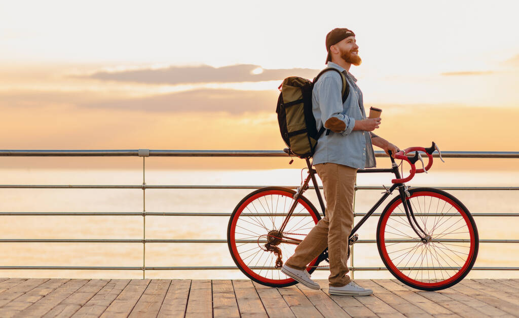 όμορφος hipster στυλ γενειοφόρος άνδρας με σακίδιο φορώντας denim πουκάμισο και καπέλο με ποδήλατο το πρωί ανατολή δίπλα στη θάλασσα πίνοντας καφέ, υγιή ενεργό τρόπο ζωής ταξιδιώτη backpacker - Φωτογραφία, εικόνα