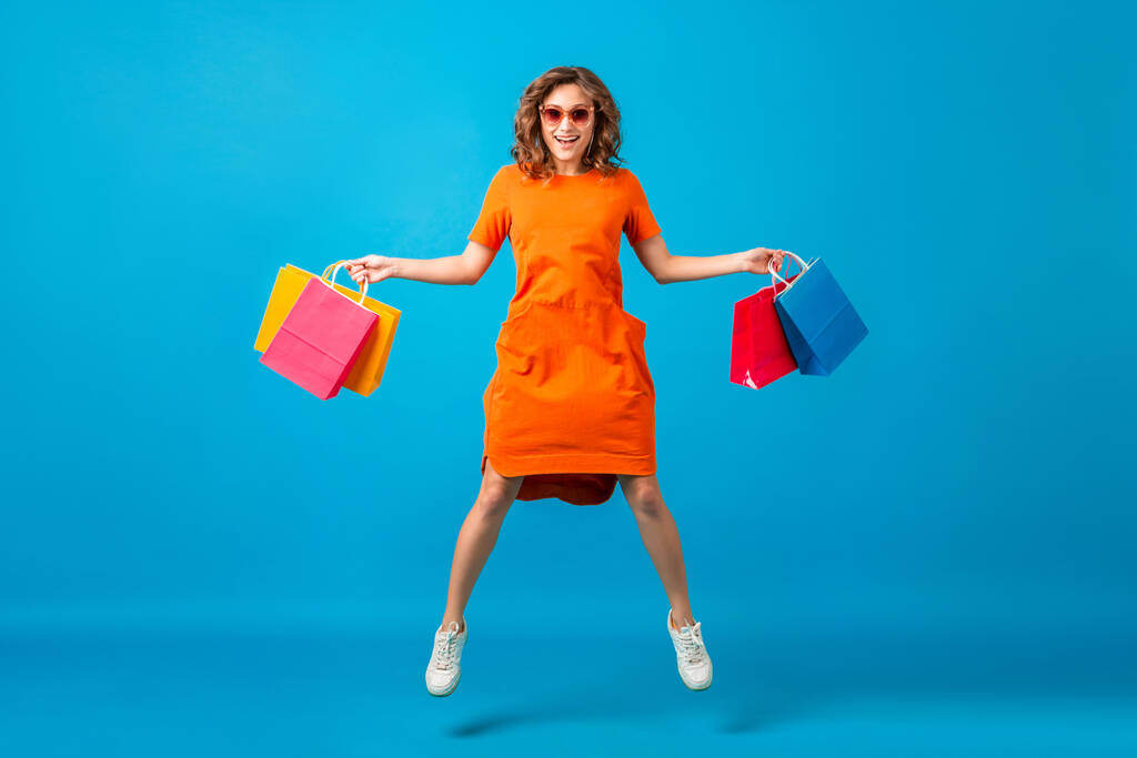 приваблива щаслива усміхнена стильна жінка шопоголіка в помаранчевій модній сукні, стрибає, тримає сумки на синьому студійному фоні ізольовані, барвисті, продаються схвильовані, весняні літні модні тенденції
 - Фото, зображення